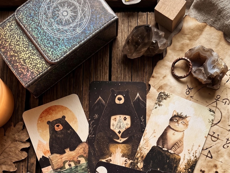 May include: A rectangular holographic storage box with a circular star design, three illustrated cards featuring a bear on a log, a bear with a geometric chest symbol, and a cat, a brown ring, and two raw crystal stones on a wooden surface.