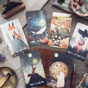 May include: A collection of seven tarot cards featuring illustrated scenes including a fox, a whale, a turtle, a raven, and a polar bear. The cards are labeled Seven of Wands, Ace of Cups, Nine of Pentacles, Three of Swords, The Magician, and Wheel of Fortune.