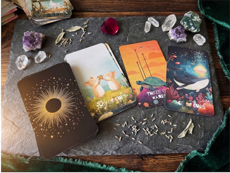May include: A tarot deck featuring a black card with a gold sunburst, a Two of Cups card with two mice, a Three of Wands card with a turtle, and an Ace of Cups card with a whale. The cards are surrounded by scattered crystals and dried lavender.