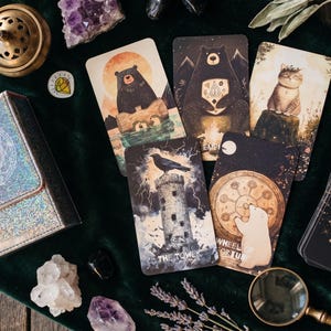 May include: A deck of tarot cards featuring animal illustrations, including a bear on a log, a bear with a campfire, a cat, a raven on a tower labeled The Tower, and a polar bear with a wheel labeled Wheel of Fortune, with a holographic box.
