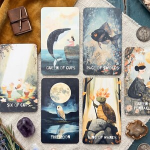 May include: A collection of six tarot cards featuring illustrations of the Queen of Cups, Page of Swords, Six of Cups, The Moon, King of Wands, and Queen of Pentacles, arranged on a textured fabric surface with various small decorative items.