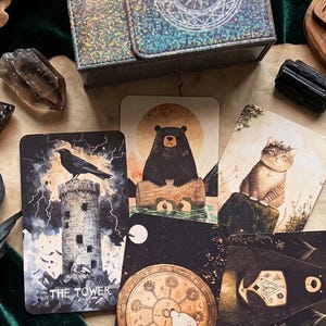 May include: A collection of illustrated tarot cards featuring a black crow on a stone tower labeled The Tower, a black bear on a log, and a tabby cat. The cards are arranged with a dark box, crystals, and wooden runes, showcasing detailed artwork.