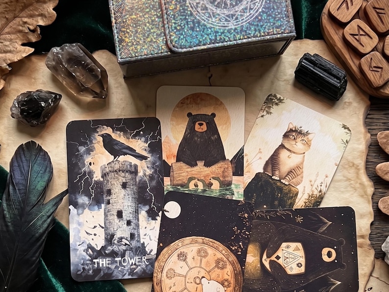 May include: A collection of illustrated tarot cards featuring a black crow on a stone tower labeled The Tower, a black bear on a log, and a tabby cat. The cards are arranged with a dark box, crystals, and wooden runes, showcasing detailed artwork.