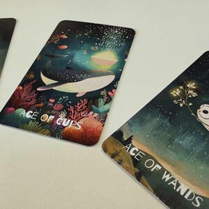 May include: Three rectangular tarot cards featuring artistic illustrations. The left card shows a fish and a sword with the text ACE OF SWORDS. The middle card depicts a whale in an underwater scene with the text ACE OF CUPS. The right card shows an owl with the text ACE OF WANDS.