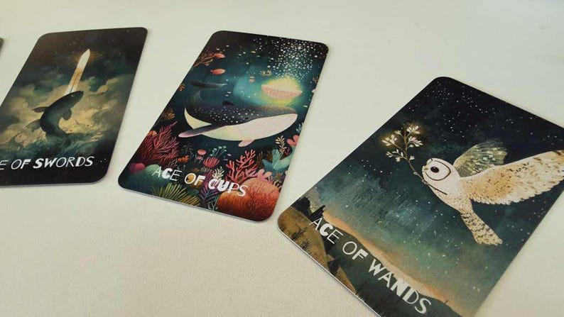 May include: Three rectangular tarot cards featuring artistic illustrations. The left card shows a fish and a sword with the text ACE OF SWORDS. The middle card depicts a whale in an underwater scene with the text ACE OF CUPS. The right card shows an owl with the text ACE OF WANDS.
