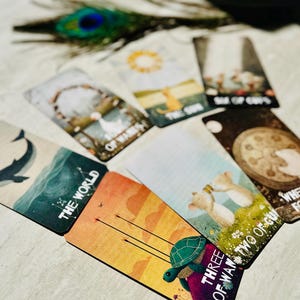 May include: A collection of tarot cards spread out, featuring illustrated designs including a whale labeled The World, a turtle labeled Three of Wands, two mice labeled Two of Cups, a hand on a wheel labeled Wheel of Fortune, and The Sun.