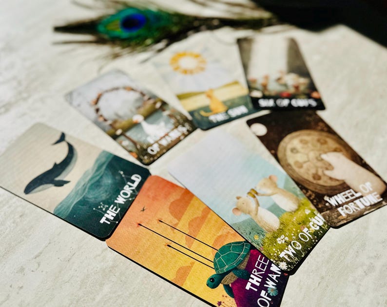 May include: A collection of tarot cards spread out, featuring illustrated designs including a whale labeled The World, a turtle labeled Three of Wands, two mice labeled Two of Cups, a hand on a wheel labeled Wheel of Fortune, and The Sun.