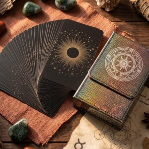 May include: A deck of black tarot cards fanned out, featuring a gold sunburst design on the back, alongside a matching iridescent holographic storage box with a circular geometric emblem printed on the front cover in a white line art style.