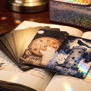 May include: A deck of tarot cards fanned out on an open book, featuring The Tower card with a black bird on a stone tower and the Wheel of Fortune card with a polar bear. The cards have dark backgrounds with gold and holographic accents.