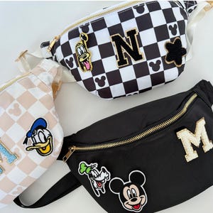 May include: Three fanny packs in a variety of patterns and colors. One is black with a gold zipper and the letter "M". Another is checkered black and white with the letter "N". The third is light pink and white checkered with the letter "M".