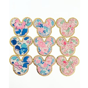 May include: Nine floral Mickey Mouse head patches. Each patch features a gold glitter border and a colorful floral pattern in shades of pink, blue, and white. The patches are arranged on a white background.
