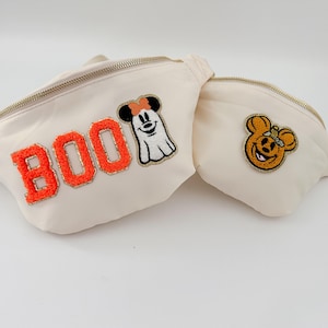 May include: Two off-white fanny packs with gold zippers. One has the word "BOO" in orange letters with a ghost-like Mickey Mouse patch. The other has a Mickey Mouse pumpkin patch. Perfect for Halloween.