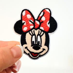 May include: Embroidered patch featuring Minnie Mouse's face. The patch has a black outline, black ears, and a red bow with white polka dots. The face is beige with black eyes, a black nose, and a red tongue.