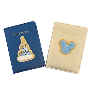 Op de afbeelding: Twee paspoorthouders. Eén is marineblauw met een kasteelontwerp en het woord "PASSPORT" in goud. De andere is beige met een Mickey Mouse-ontwerp en het woord "PASSPORT" in goud.