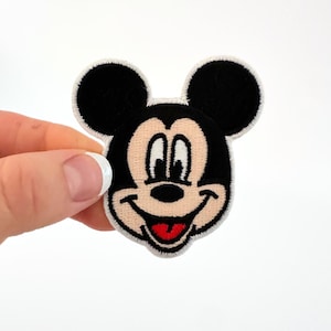 Peut inclure: Un écusson Mickey Mouse représentant un visage souriant avec des oreilles noires, une tête noire et un visage beige. L'écusson a une langue rouge et des yeux blancs. L'écusson est bordé de blanc et a une bordure cousue.