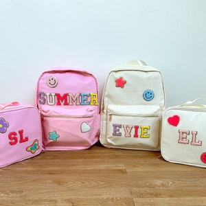 Peut inclure: Ensemble de sacs à dos et de sacs à lunch roses et crème. Les articles roses affichent le mot "SUMMER" et les initiales "SL". Les articles crème présentent le nom "EVIE" et les initiales "EL". Chaque sac est orné d'écussons colorés.