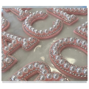 Może przedstawiać: Zbliżenie kilku różowych filcowych liter, każda obramowana małymi, błyszczącymi kryształkami i ozdobiona rzędem białych sztucznych pereł. Litery ułożone są na białej powierzchni, tworząc dekoracyjną ekspozycję.