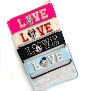 Könnte beinhalten: Vier farbenfrohe Reißverschlusstaschen mit dem Wort "LOVE" in Glitzerbuchstaben. Jede Tasche zeigt einen anderen Disney-Charakter: Daisy Duck, Minnie Mouse, Mickey Mouse und Donald Duck. Die Taschen sind rosa, cremefarben, schwarz und hellblau.