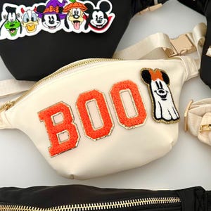 Può includere: Marsupio color crema con la scritta "BOO" in arancione, bordata d'oro. Una toppa a forma di fantasma di Topolino è a destra. Sono visibili altri marsupi neri con toppe di personaggi di Halloween.
