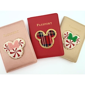 May include: Three passport holders in pink, red, and gold. Each has a decorative Mickey Mouse-shaped applique. The pink holder has a pink bow and red and white candy accents. The red holder has a plaid applique. The gold holder has a green bow and candy accents.