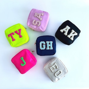 May include: A selection of small, square pouches in a range of colours, each featuring embroidered initials. Colours include pink, black, neon green, navy, hot pink, and cream. The initials are in contrasting colours and textures, adding a personalised touch to these accessories.