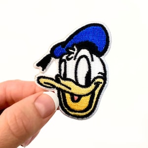 Peut inclure: Écusson brodé représentant le visage souriant de Donald Duck. Le patch a un visage blanc, un bec jaune et un chapeau de marin bleu. Il est bordé de noir et a une bordure cousue.