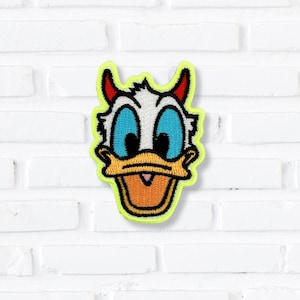 May include: An embroidered patch featuring the head of a cartoon duck with white feathers, large blue eyes, and an orange beak. The character has small red horns on top of its head and is outlined by a bright neon yellow border.