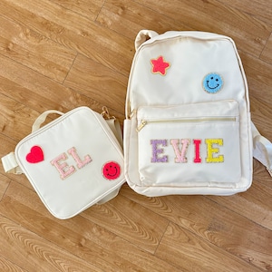 May include: Cream-coloured backpack and lunch bag set. The backpack features a gold zip and the name "EVIE" in colourful, glittery letters. The lunch bag has the initials "EL" in glittery letters. Both bags have decorative patches, including a star, heart, and smiley face.