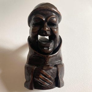Könnte beinhalten: Dunkelbrauner, geschnitzter Nussknacker aus Holz in Form einer lächelnden Figur mit Hut und Robe. Der Mund der Figur ist geöffnet und gibt den Nussknackmechanismus frei. Die Hände sind vor der Brust gefaltet.