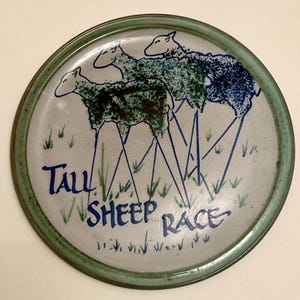 May include: A round ceramic plate with a green rim. The plate features a whimsical design of three sheep with long, thin blue legs, racing across a field of grass. The words "Tall Sheep Race" are written in blue.
