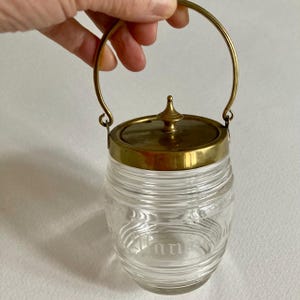 Könnte beinhalten: Ein klares Glasgefäß mit Messingdeckel und Griff. Das Gefäß hat ein geripptes Design und das Wort "mm" ist auf der Vorderseite eingraviert. Der Deckel ist mit einem dekorativen Knauf versehen. Der Griff ist gewölbt und mit kleinen Schlaufen am Gefäß befestigt.