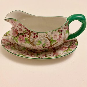 May include: A floral gravy boat and saucer set. The boat has a green handle and rim, and a spout for pouring. The set is decorated with pink flowers, red berries, and grey branches on a white background. The saucer has a matching floral pattern.