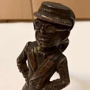 May include: A cast iron figurine of a man wearing a hat and glasses. The figurine is painted in a dark brown colour and has a distressed finish. The man is standing with his hands on his hips and has a serious expression on his face.
