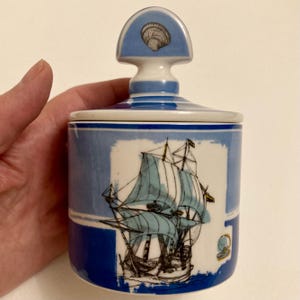 May include: A blue and white ceramic jar with a lid featuring a seashell design. The jar's body displays a detailed illustration of a sailing ship with blue sails. The jar is held by a person.