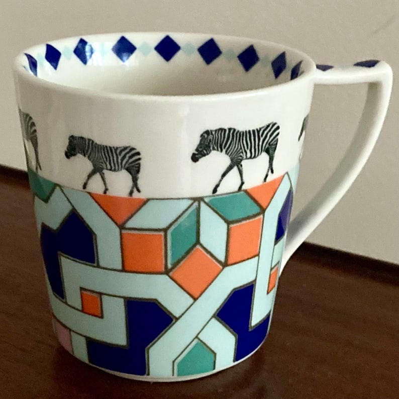 Puede incluir: Taza de cer&aacute;mica blanca con un patr&oacute;n geom&eacute;trico en azul, naranja y verde azulado. La parte superior de la taza presenta ilustraciones de cebras en blanco y negro y formas de diamantes azules. La taza tiene un asa blanca.