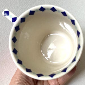 Puede incluir: Una taza de t&eacute; de cer&aacute;mica blanca con asa, con un dise&ntilde;o de borde de formas de diamantes azules y celeste alternadas. El interior de la taza es liso y brillante, reflejando la luz. El asa tambi&eacute;n tiene un patr&oacute;n de diamantes.