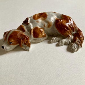 以下が含まれることがあります： 休息姿勢の犬の陶器製置物。犬は白い毛並みで、頭と耳に茶色の斑点と模様があります。置物は青い目を持ち、横たわっています。