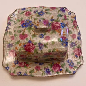 Peut inclure: Un beurrier vintage avec couvercle, orné d'un motif floral de roses roses, de fleurs bleues et violettes et de feuilles vertes sur fond crème. Le plat est de forme carrée avec un bord festonné.