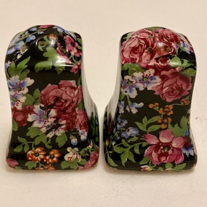 Puede incluir: Un par de saleros y pimenteros de cerámica con fondo negro y estampado floral. El estampado floral incluye flores rosas, rojas, azules y naranjas con hojas verdes. Los saleros y pimenteros tienen forma rectangular.