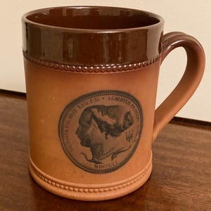 Może przedstawiać: Brązowy ceramiczny kubek z uchwytem i dekoracyjnym obramowaniem. Posiada okrągły emblemat z portretem profilowym. Kubek ma teksturowaną podstawę i ciemnobrązową krawędź.
