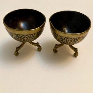 A pair of vintage, Indian, brass covered coconut shell small bowls, with brass scrolled tripod feet. One of the cups has a dent on the side.