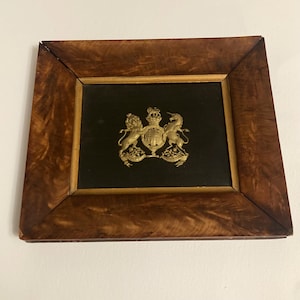 Könnte beinhalten: Ein gerahmtes Emblem mit goldenem Wappen auf schwarzem Hintergrund. Der Holzrahmen hat ein braunes, wirbelndes Maserungsmuster. Das Emblem enthält eine Krone, Löwen und ein Einhorn.