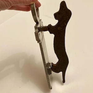 May include: A vintage metal door knocker featuring a silhouette of a dog. The knocker is made of a dark, weathered metal and has a rectangular backplate. The dog silhouette is attached to the backplate with a hinge.