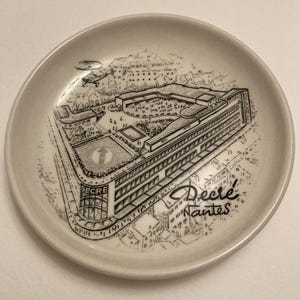 Puede incluir: Un plato pequeño de cerámica blanquecina con una ilustración en blanco y negro de un edificio, con el texto "Decré Nantes". El plato presenta un dibujo arquitectónico detallado del edificio y sus alrededores.