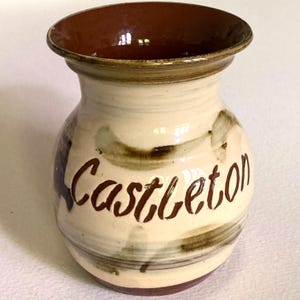 May include: A ceramic vase with a cream-colored body and a dark brown interior rim. The exterior features hand-painted dark brown brushstrokes and the word Castleton written in a cursive script across the front. The base is also dark brown.