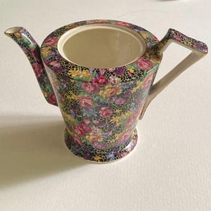 May include: A ceramic teapot featuring a vibrant floral pattern of pink, yellow, and purple roses set against a black honeycomb background. The teapot has a wide circular opening, a curved spout, and an angular handle with gold-toned trim.