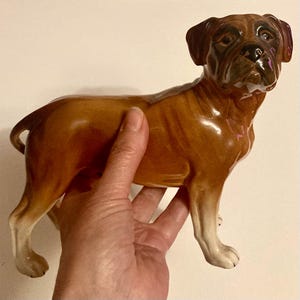 Puede incluir: Figura de perro Boxer de cerámica en posición de pie. El perro tiene un acabado brillante con colores marrón y canela. La figura está sujeta por una mano.