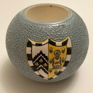 May include: A small, round, gray ceramic container with a textured surface. It features a decorative shield with black, white, and yellow details. The interior is a light beige color.