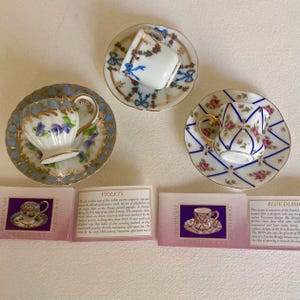 May include: Four miniature teacups and saucers with floral and geometric designs. The teacups are white with gold trim and the saucers have blue and gold accents. The teacups are arranged in a row on a white surface. The text on the cards reads "Violets", "Blue Diamond", and "Certificate".