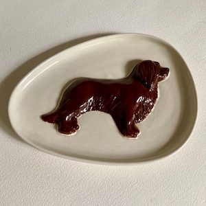 May include: A white ceramic oval dish with a brown ceramic dog sculpture in relief. The dog is standing with its head turned to the right.